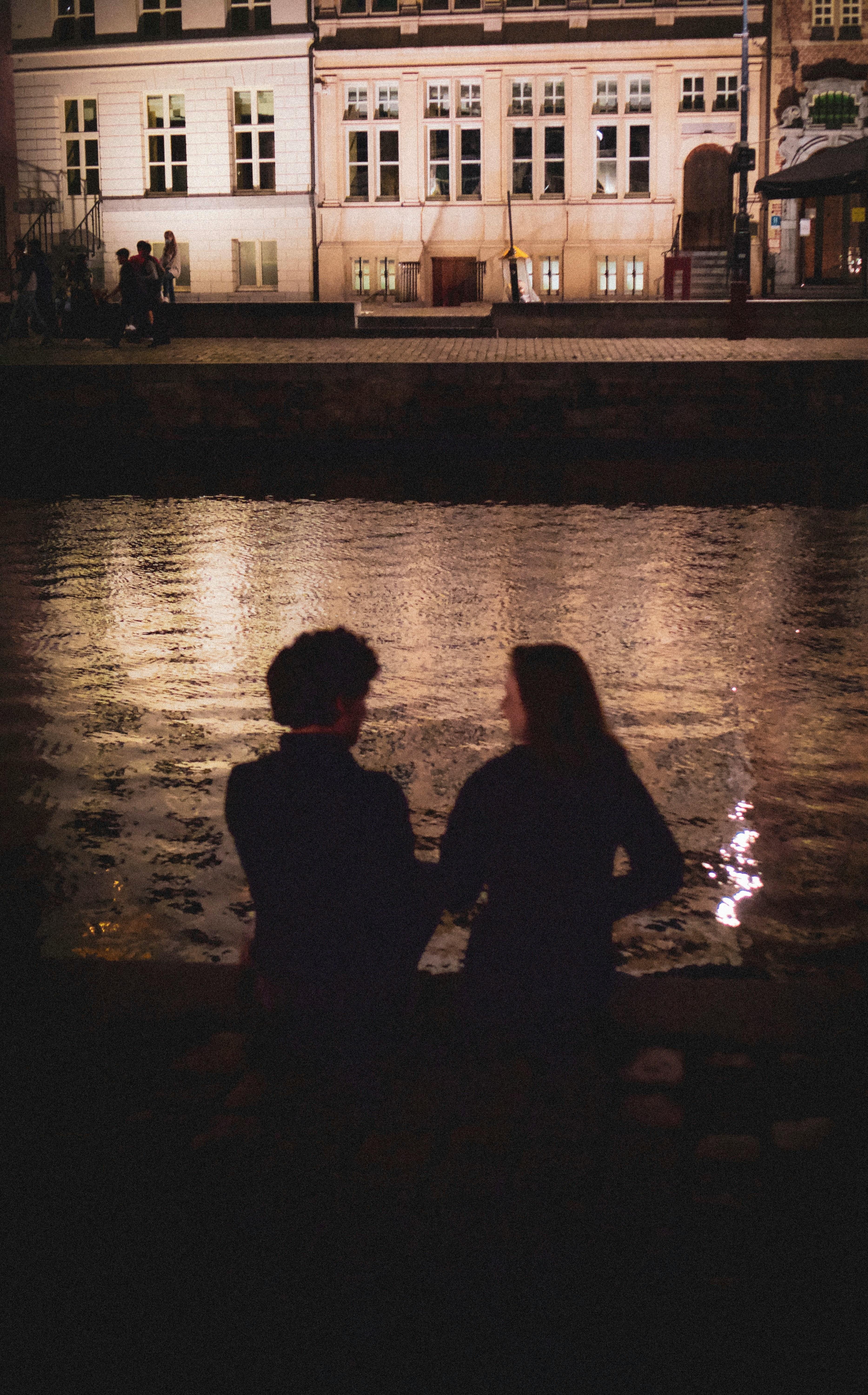 Couple silhouetted by a canal at night, with illuminated buildings in the background